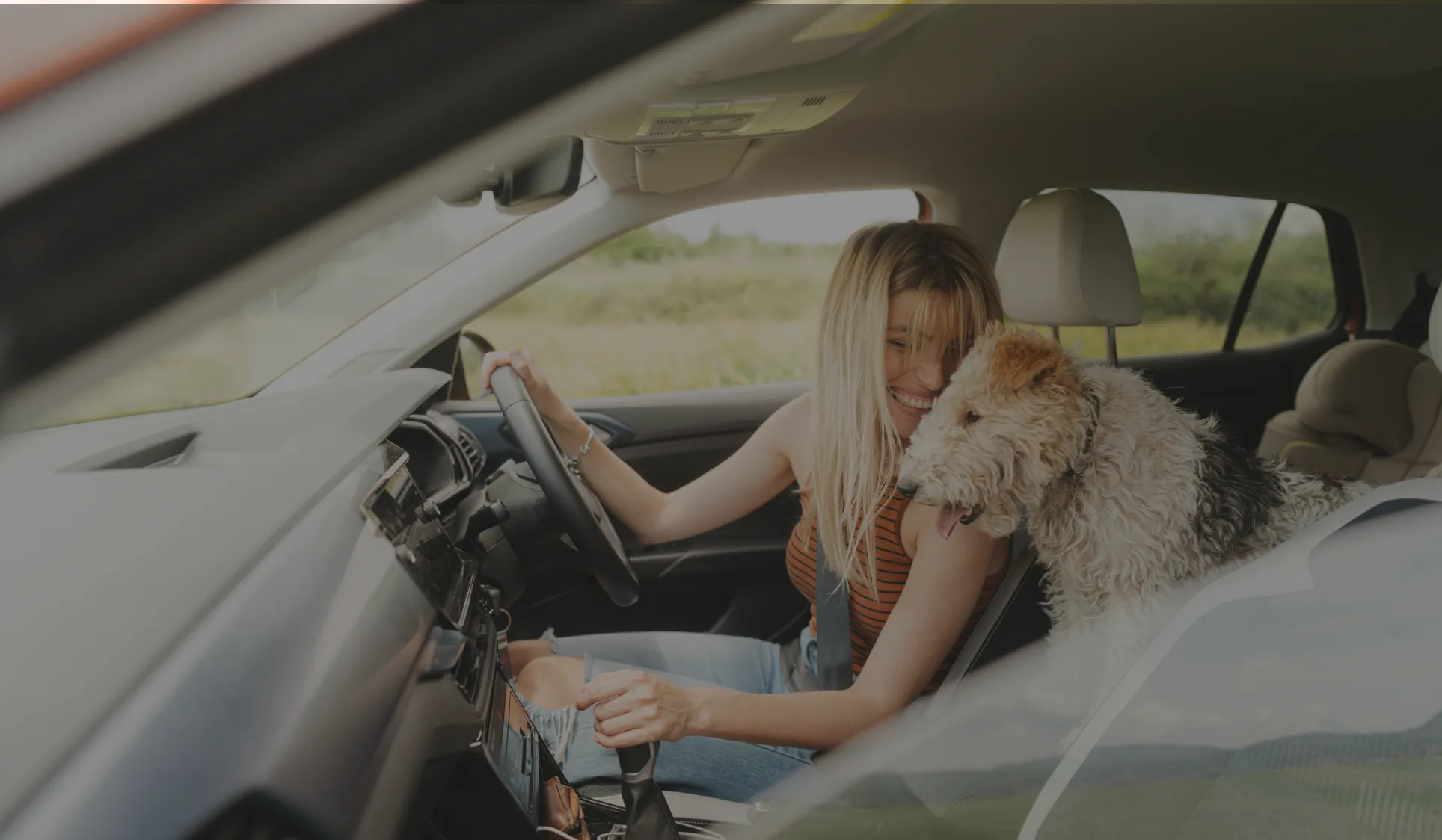 A woman sits in the driver seat of her car smiling at her dog between the seats.